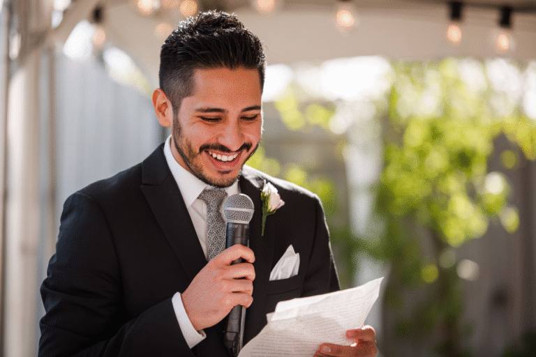 A man in a suit and tie holds a microphone and reads from a sheet of paper while smiling, standing outdoors under string lights.