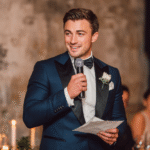 A man in a blue tuxedo holds a microphone and a piece of paper, speaking at an indoor event with candles and blurred guests in the background.
