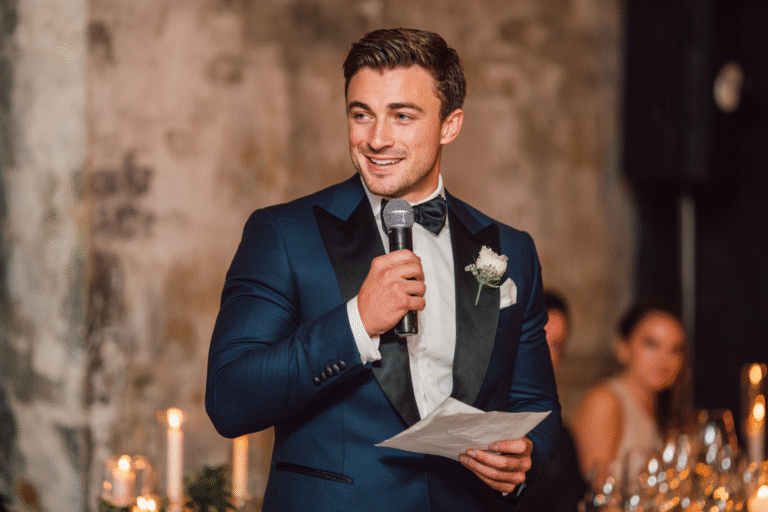 A man in a blue tuxedo holds a microphone and a piece of paper, speaking at an indoor event with candles and blurred guests in the background.