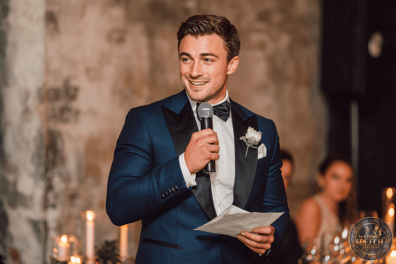 A man in a blue tuxedo holds a microphone and a piece of paper, speaking at an indoor event with candles and blurred guests in the background.