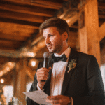 A man in a tuxedo holds a microphone and a piece of paper, speaking at an indoor event with exposed wooden beams and string lights.