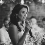 A woman in a white dress holds a microphone and speaks to a seated group of people at an indoor event.