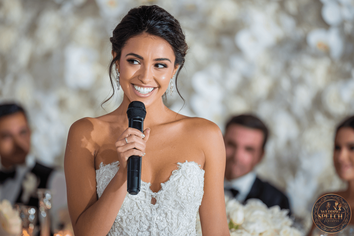 A bride in a strapless white wedding dress smiles while holding a microphone, with guests seated and blurred in the background.