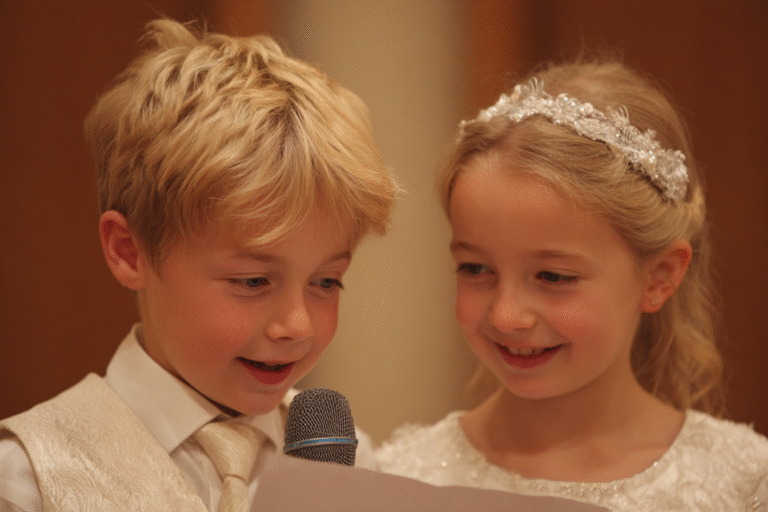 Two young children dressed formally, with a boy holding a microphone and a sheet of paper, stand closely together and appear to be speaking or reading aloud.