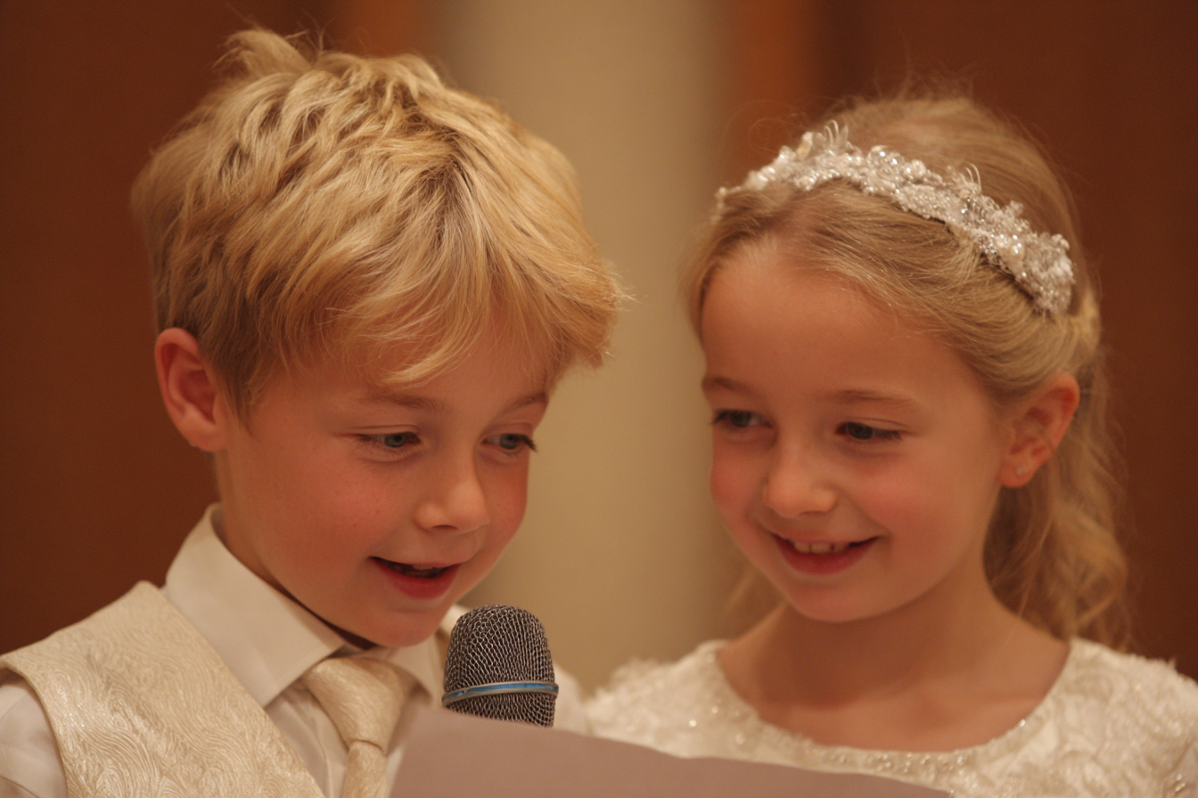 Two young children dressed formally, with a boy holding a microphone and a sheet of paper, stand closely together and appear to be speaking or reading aloud.