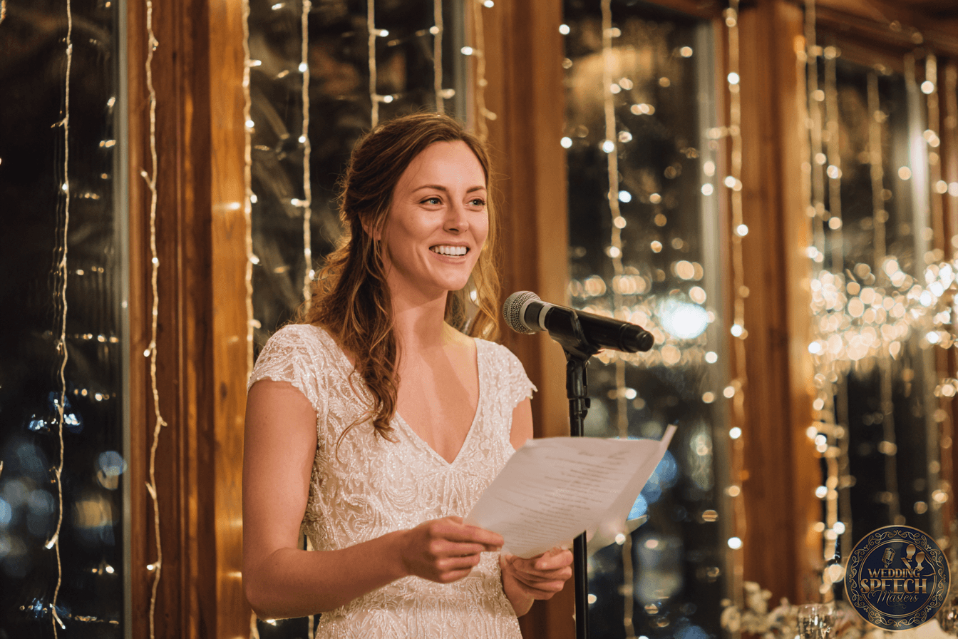 A woman in a white dress stands at a microphone, holding a sheet of paper, speaking in front of large windows decorated with string lights.