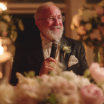 An older man in a suit and bow tie holds a microphone and smiles while giving a speech at a formal event, surrounded by flowers and soft candlelight.