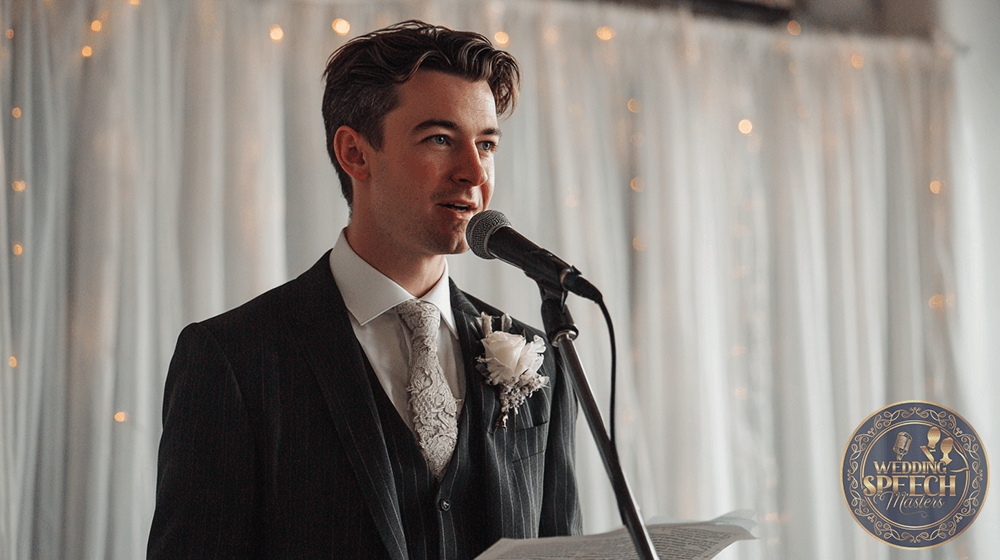 A man in a suit with a boutonnière speaks into a microphone, holding papers, with white drapes and string lights in the background.