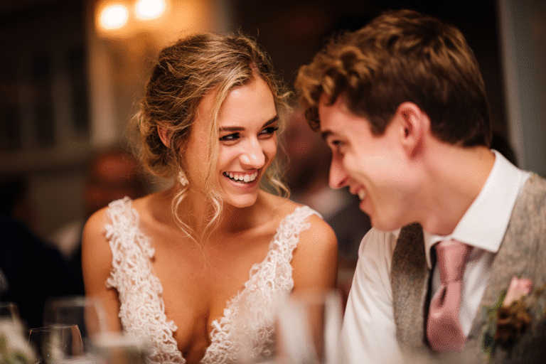 A smiling couple dressed in formal attire sits close together at a well-lit indoor event, appearing to enjoy a happy conversation.