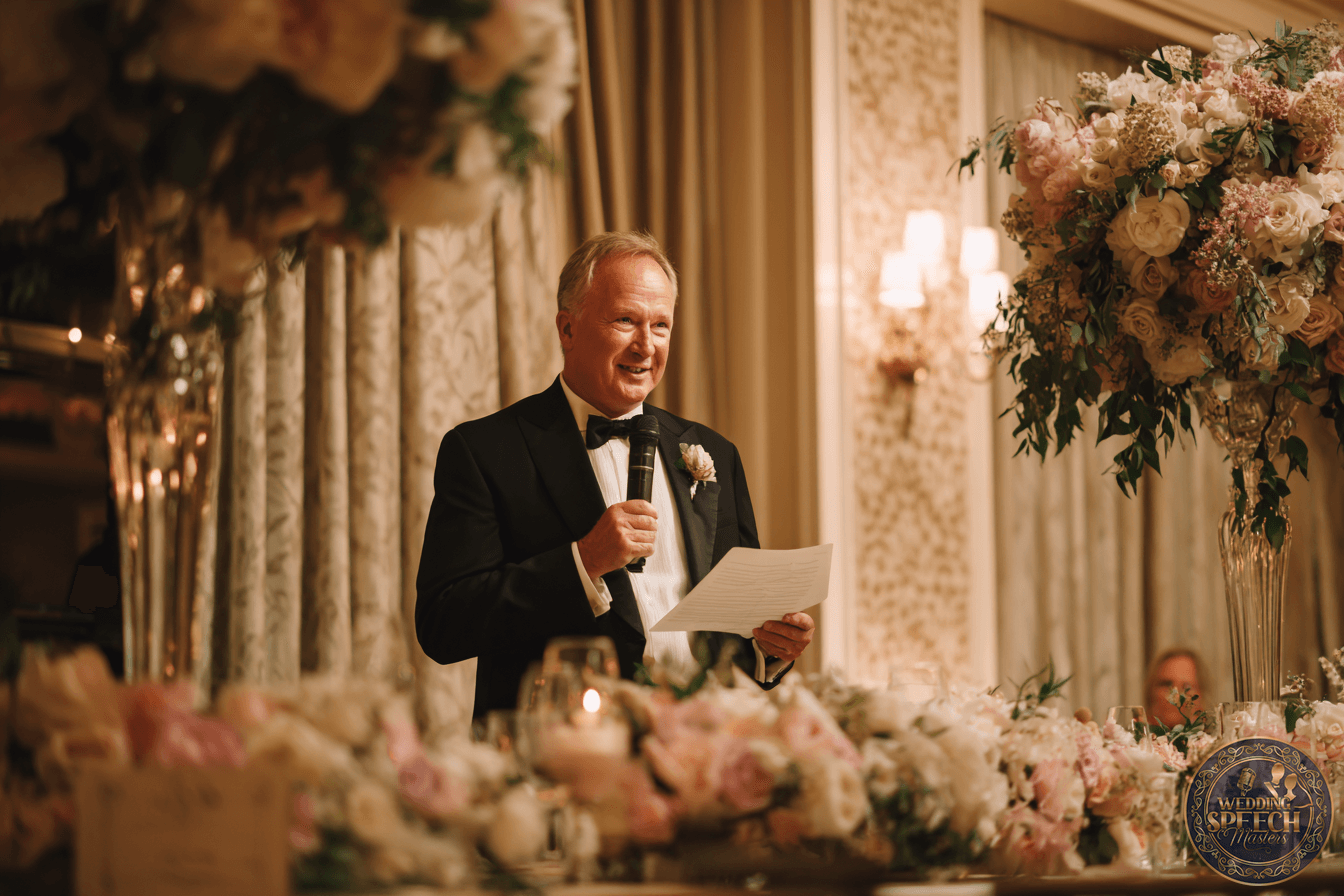 An older man in a tuxedo speaks into a microphone while holding a paper, standing behind a table with floral arrangements at a formal event.