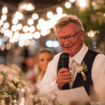 An older man in a suit and tie holds a microphone and delivers a heartfelt Father of the Bride Wedding Speech at a formal event, surrounded by flowers and soft, warm lighting.