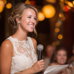 A woman in a white lace dress holds a microphone and a paper, smiling as she speaks at an indoor event with warm, blurred lights in the background.