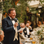 A man in a suit and bow tie stands holding a microphone and paper, smiling as he gives a speech at an outdoor formal event with seated guests.