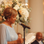 A woman in a light blue dress stands at a microphone, reading from a piece of paper, with floral arrangements and seated guests in the background.