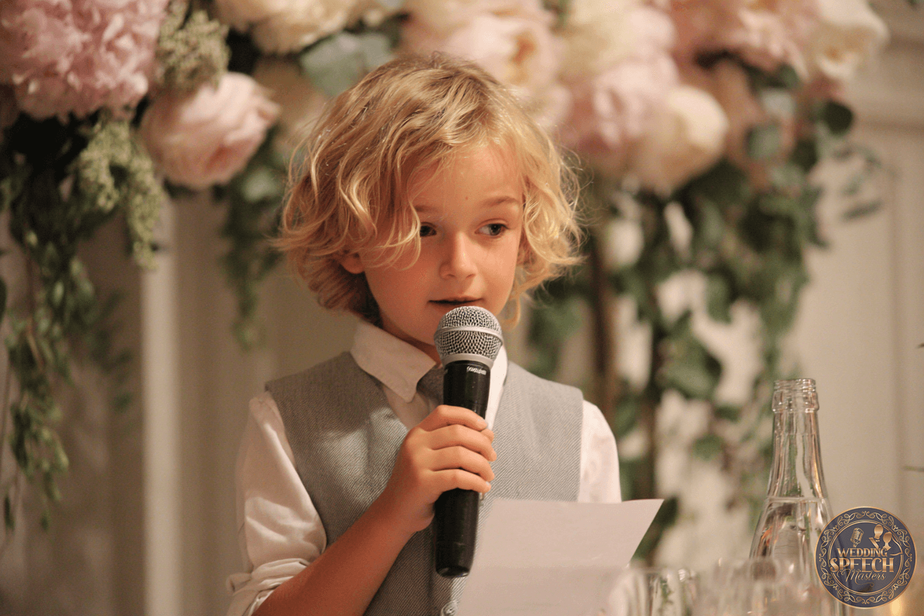 A young child with blonde hair, dressed in a white shirt and grey vest, speaks into a microphone while holding a piece of paper; floral decorations are in the background.