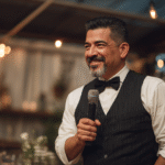 A man in a black vest and bow tie holds a microphone and smiles while speaking at an indoor event with warm lighting.