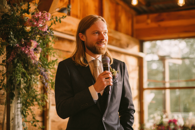 A man with long hair and a beard, dressed in a suit and tie, holds a microphone and stands indoors near floral arrangements.