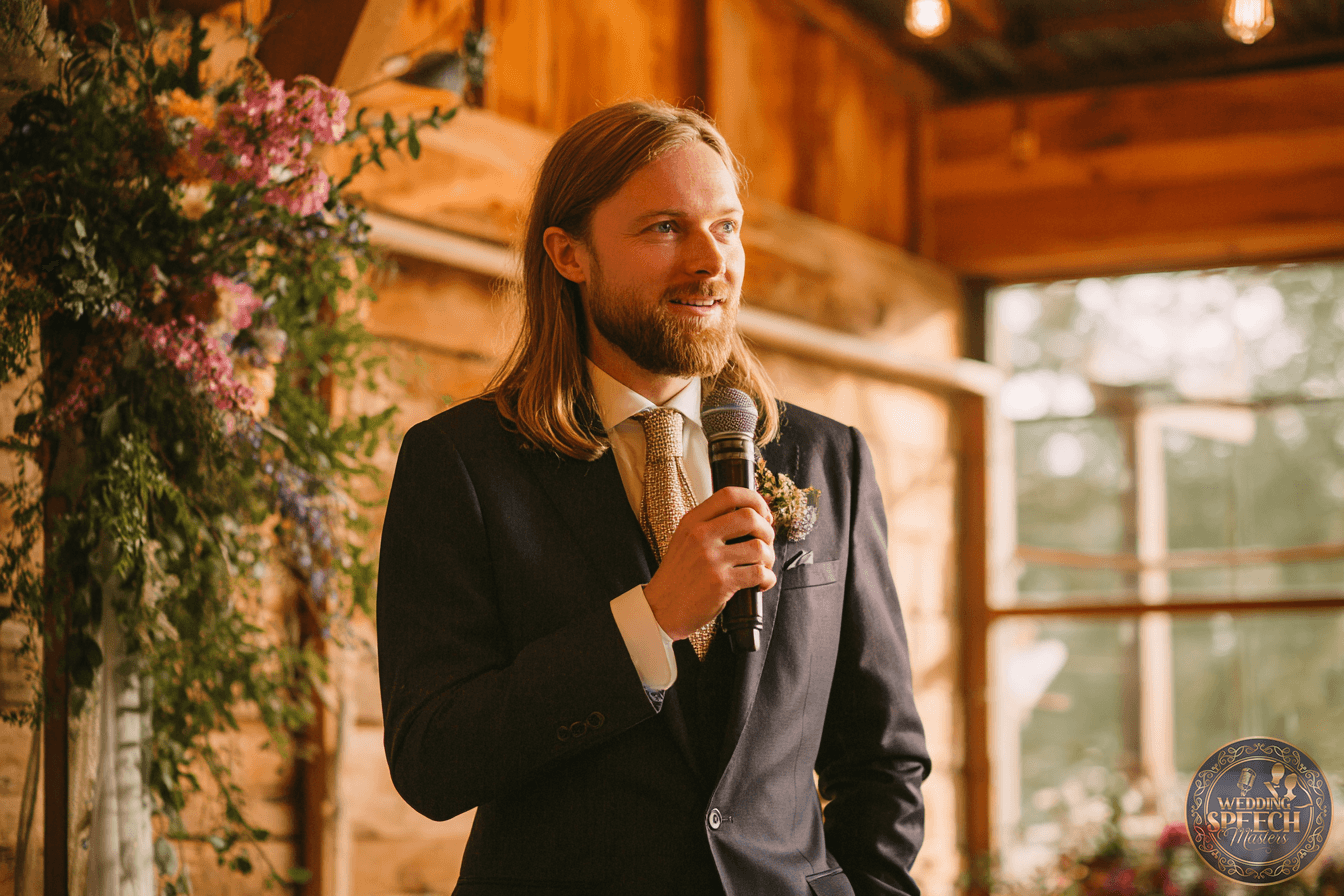 A man with long hair and a beard, dressed in a suit and tie, holds a microphone and stands indoors near floral arrangements.