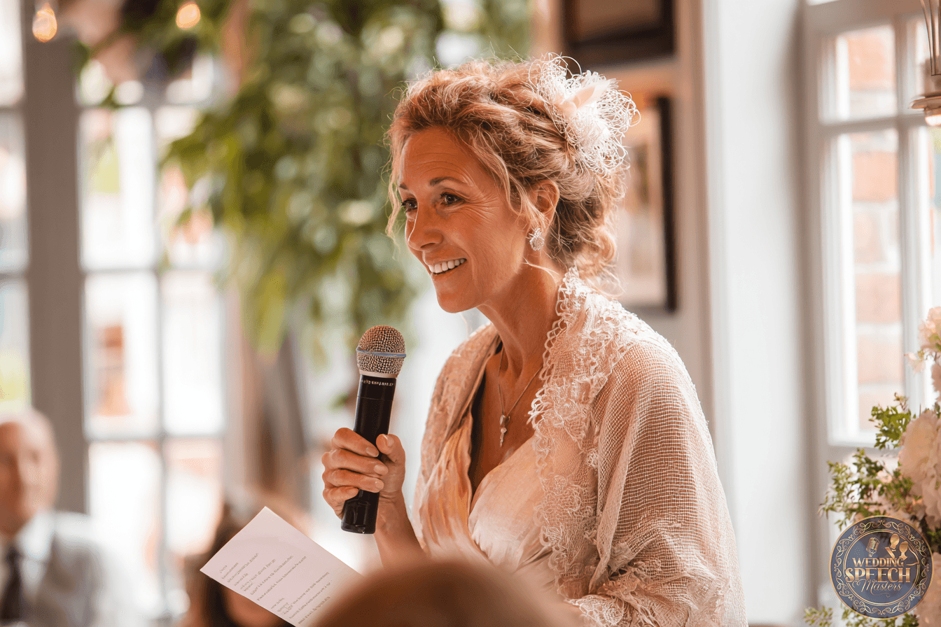 A woman in a lace shawl holds a microphone and a sheet of paper, smiling while speaking at an indoor event with natural light.