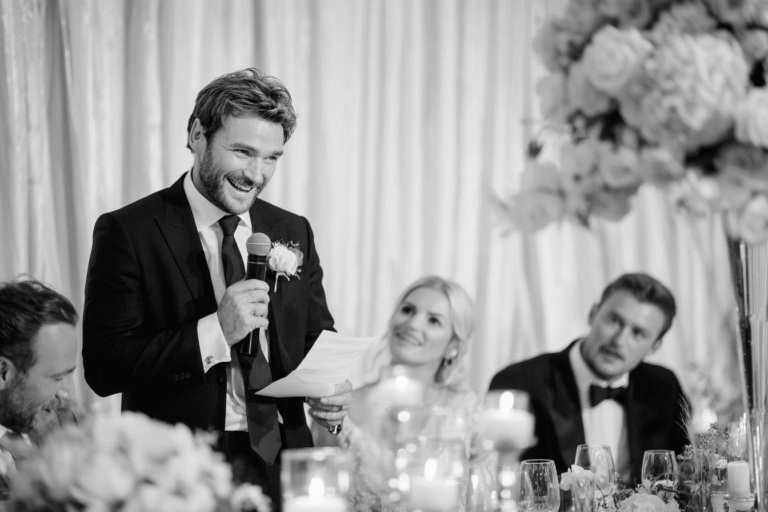 A man in a suit speaks into a microphone while holding a paper at a formal event, with seated guests and floral decorations nearby.