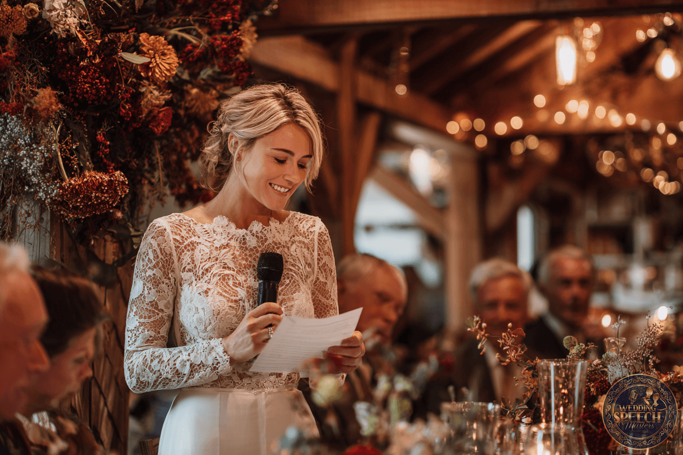A woman in a lace dress stands holding a microphone and reading from a paper, speaking to seated guests at a decorated indoor event.