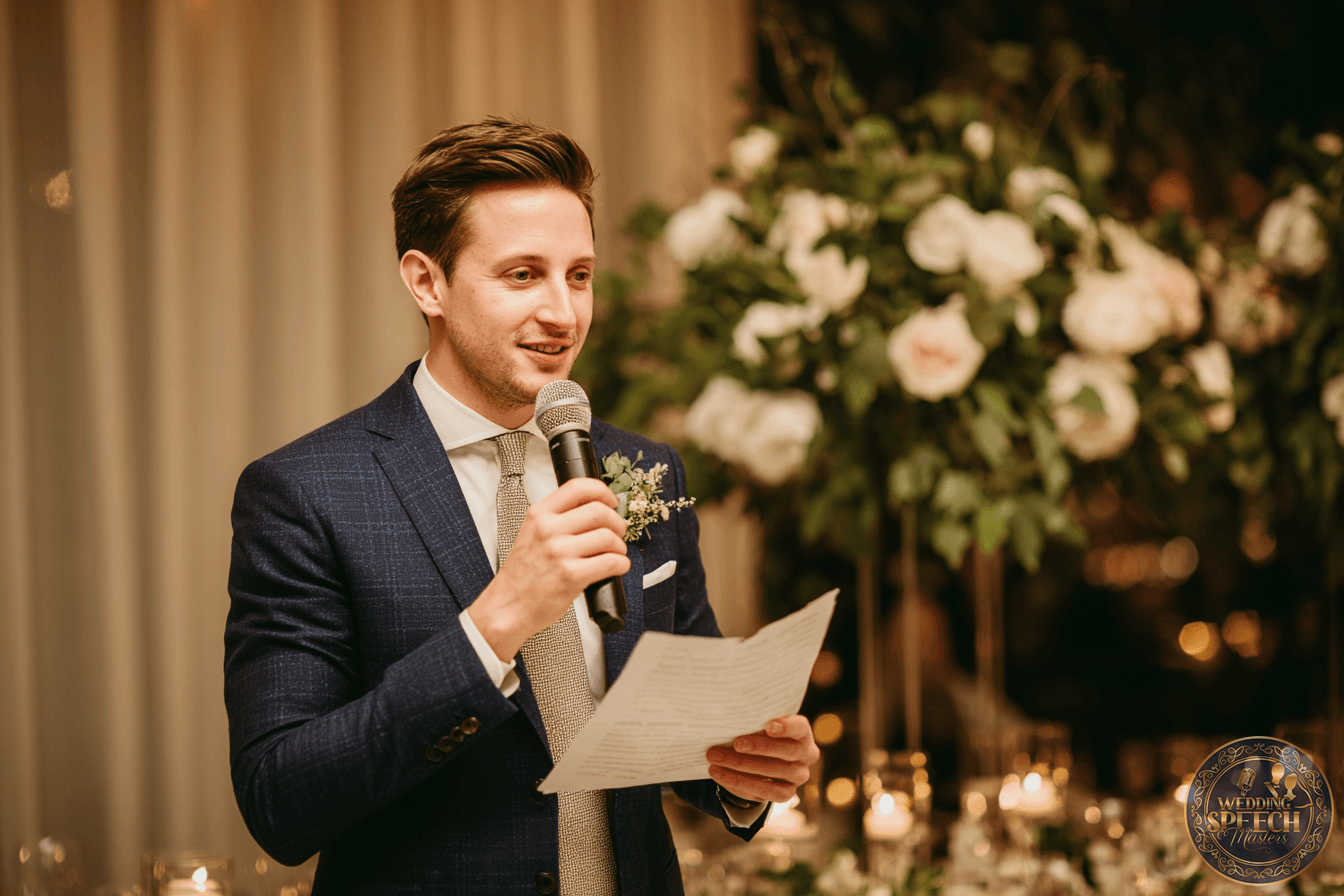 Man in a suit holding a microphone and reading from a piece of paper, standing in front of a floral arrangement at a formal event.