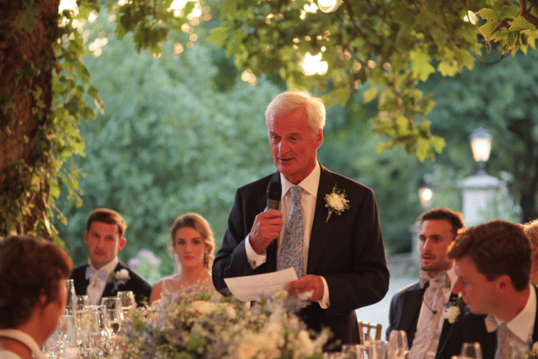 An older man in a suit gives a speech with a microphone at an outdoor event, surrounded by seated guests and greenery, with floral arrangements on the table.