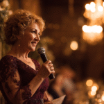 An older woman in an elegant burgundy dress holds a microphone and speaks at a formal event with warm lighting and blurred guests in the background.