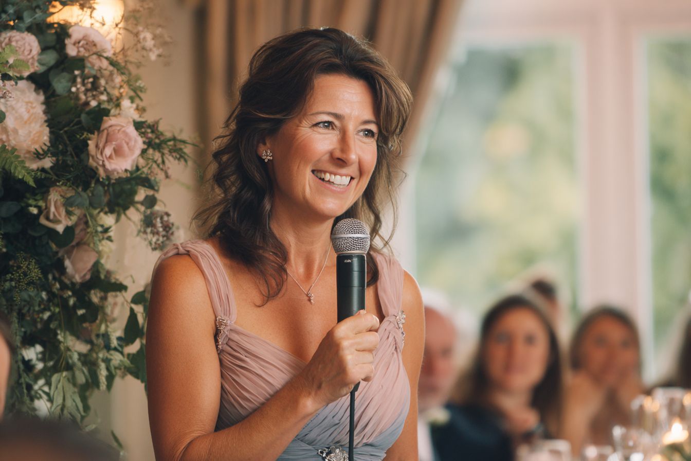 A woman in a formal dress holds a microphone and smiles while speaking at an indoor event with guests seated in the background.