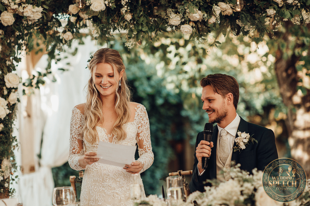 A bride in a lace wedding dress reads from a paper while a groom in a suit with a boutonniere holds a microphone, both seated under a floral arch at an outdoor wedding.