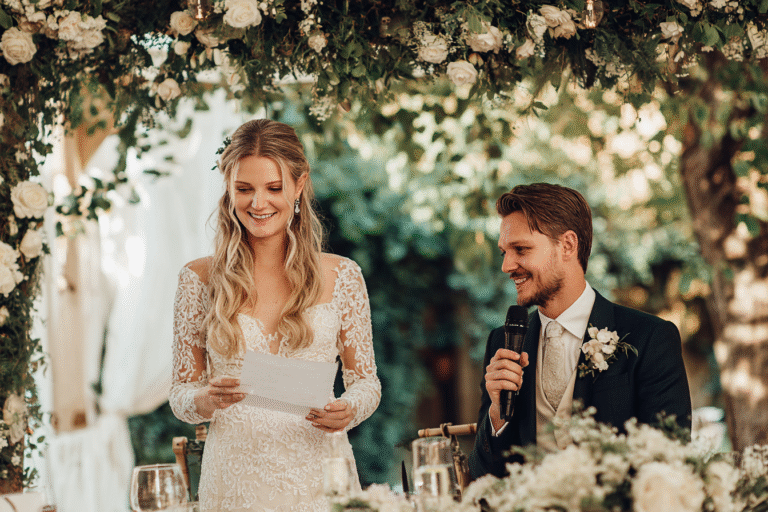 A bride in a lace wedding dress reads from a paper while a groom in a suit with a boutonniere holds a microphone, both seated under a floral arch at an outdoor wedding.