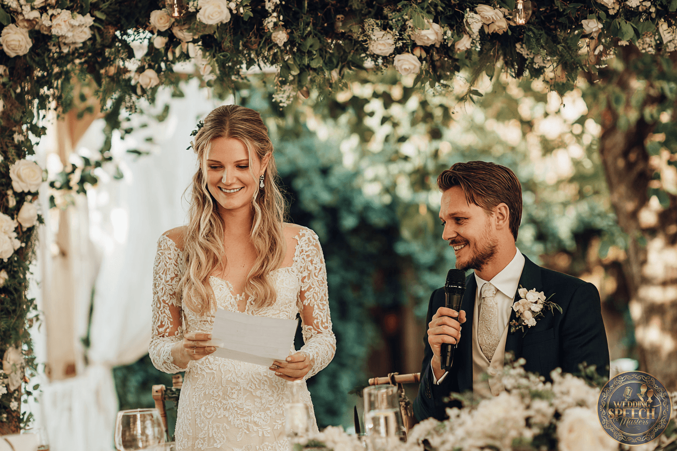 A bride in a lace wedding dress reads from a paper while a groom in a suit with a boutonniere holds a microphone, both seated under a floral arch at an outdoor wedding.