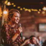 A woman in a floral dress holds a microphone and a piece of paper, speaking to an audience in a warmly lit, rustic indoor setting.
