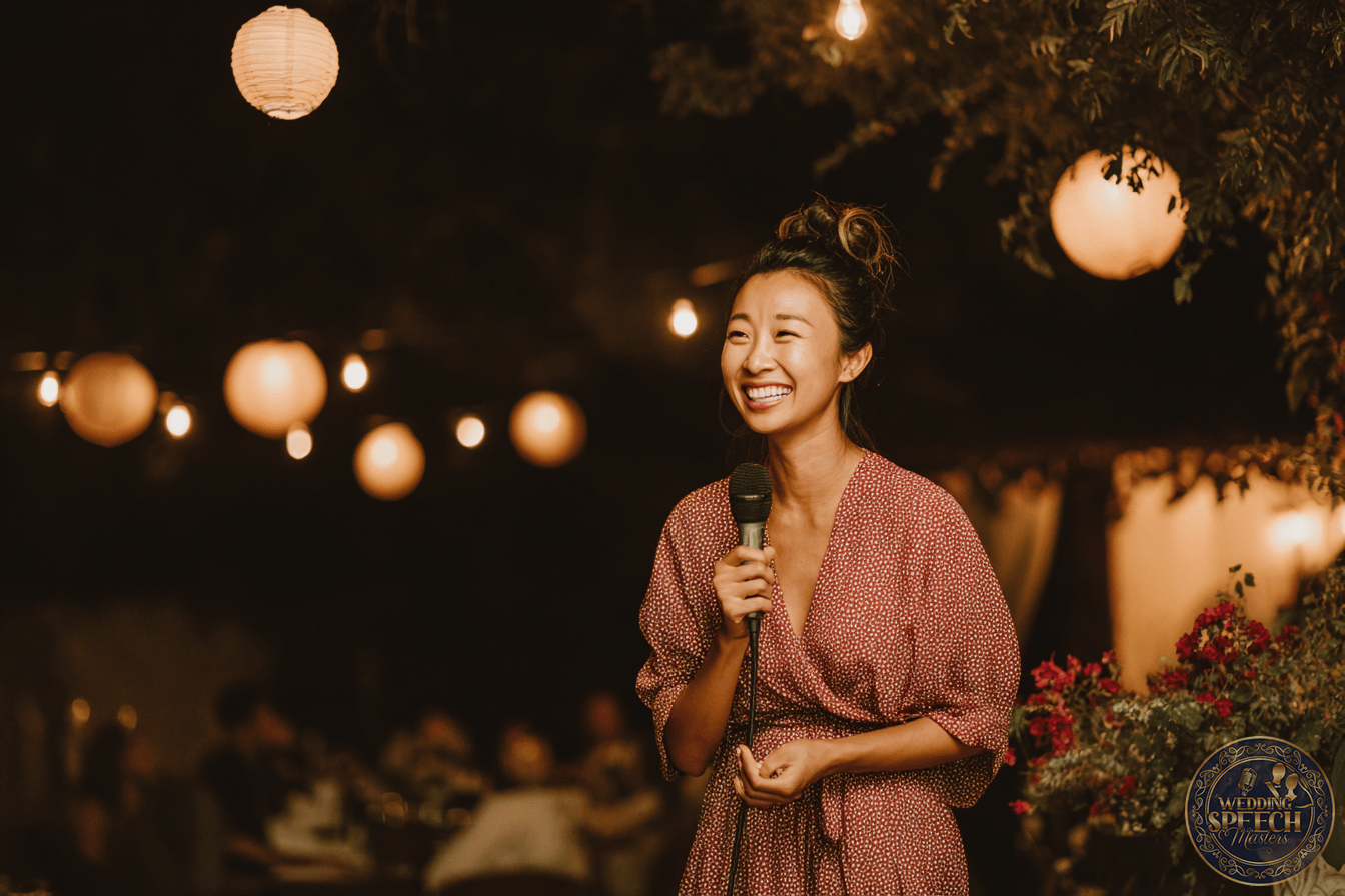 A woman in a patterned dress smiles while holding a microphone at an outdoor event with string lights and lanterns in the background.