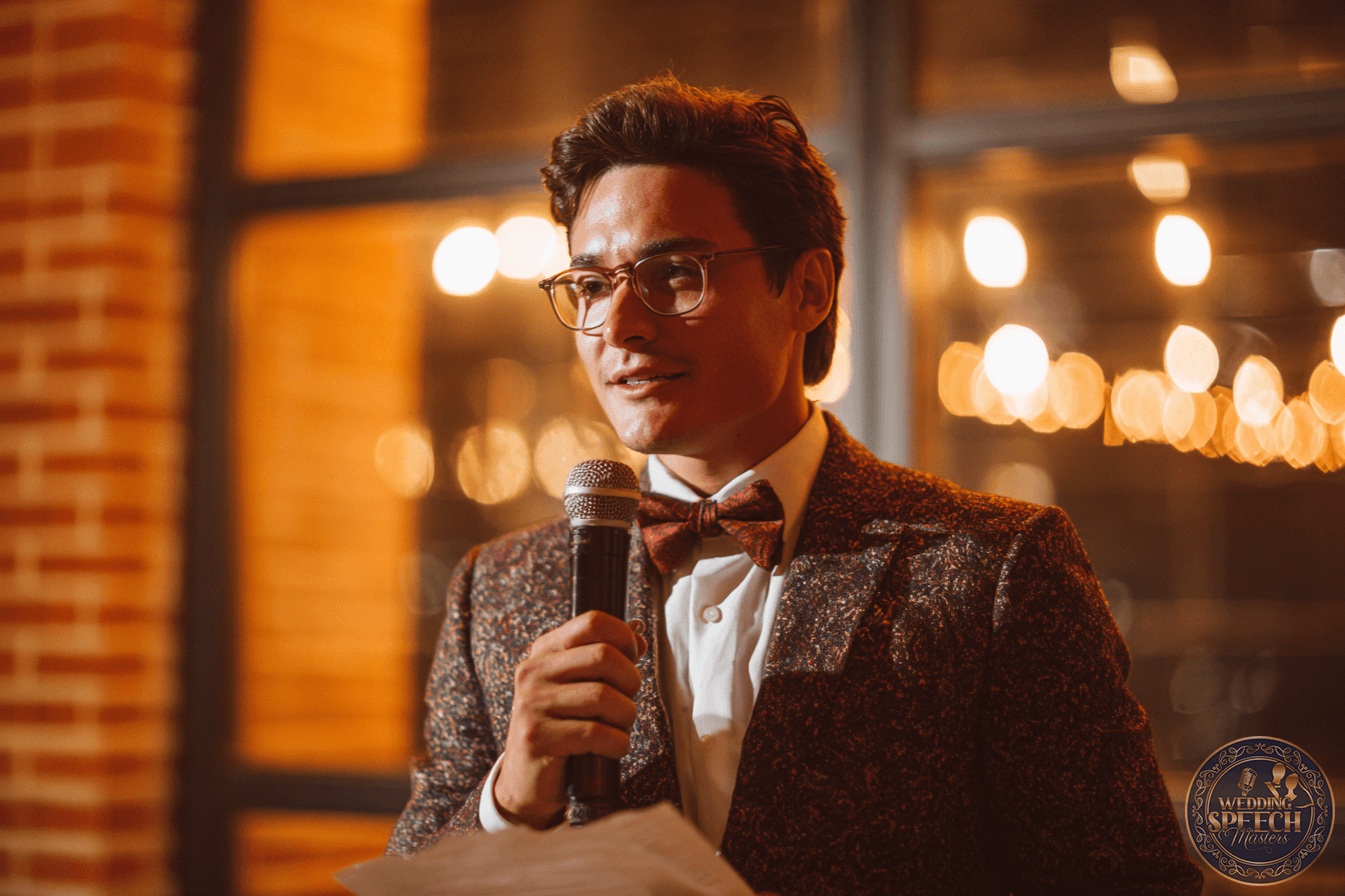A man in a patterned suit and bow tie holds a microphone and a piece of paper, speaking indoors with warm lighting in the background.