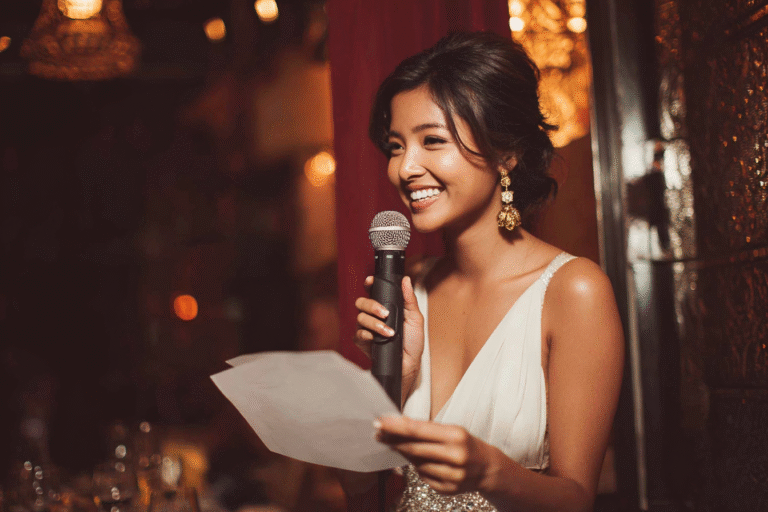 A woman in a white dress holds a microphone and a sheet of paper, smiling while speaking at an indoor event with warm lighting.