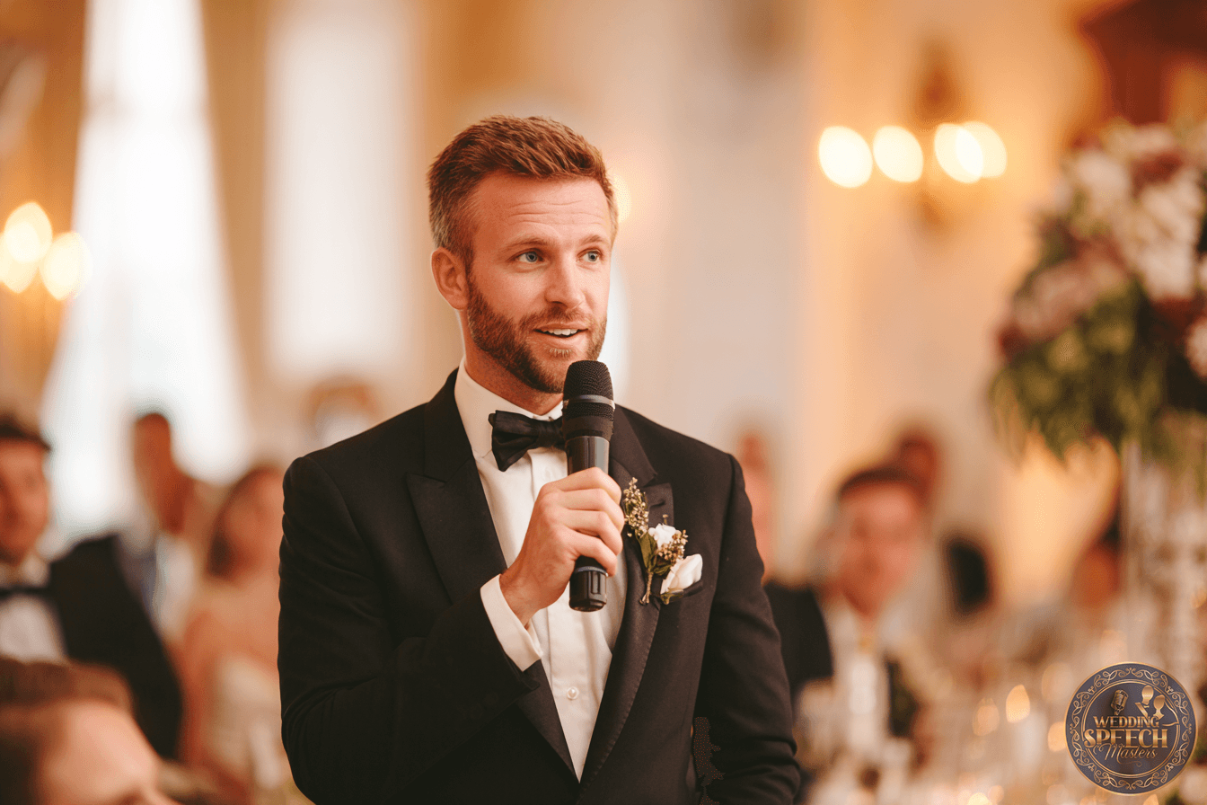 A man in a tuxedo holds a microphone, speaking at a formal indoor event with blurred guests and warm lighting in the background.